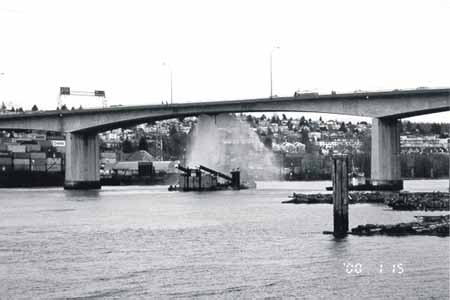The Knight Street Bridge South seen from the west immediately after the occurrence. The barge T.L. Sharpe with spuds in the cradle and pointing forward is anchored under the bridge by the sunken crane astern. Water is escaping from the broken pipe.